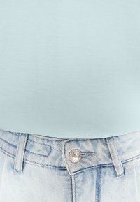 T-shirt azzurro chiaro abbinato a pantaloni corti in denim chiaro a vita alta. I dettagli notevoli includono un bottone metallico e cuciture a vista.