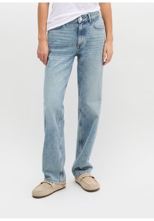 Person, die hellblaue Jeans mit geradem Bein und beige Slipper trägt, steht vor einem schlichten hellen Hintergrund.