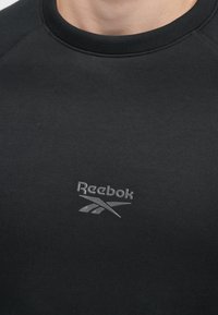 Primer plano de una persona que lleva una camiseta negra de cuello redondo con un pequeño logotipo gris de Reebok en el pecho.
