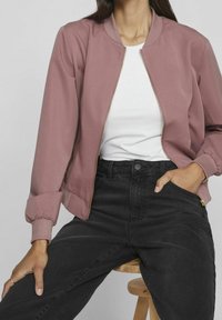 Woman wearing mauve zip-up bomber jacket, white t-shirt, and black high-waist jeans, seated on a wooden stool with one hand in pocket.