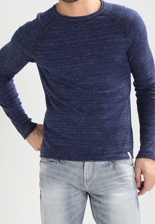 Chemise en maille texturée bleu marine à manches longues avec un col rond et des manches raglan, associée à un jean bleu clair. Petit logo discret sur l'ourlet.