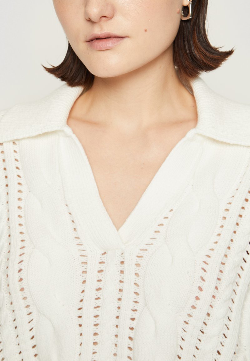 Maglione bianco lavorato a maglia con scollatura a V e colletto a coste; presenta motivi a treccia e cuciture aperte per una maggiore texture.