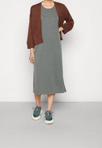 Cardigan marrone sopra un vestito midi grigio con spacchi laterali, abbinato a sneakers teal con un design testurizzato e suola chiara.
