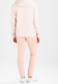 Světle růžová bunda s kapucí a elastickými manžetami, doplněná o sladěné růžové zúžené joggery a bílé tenisky na jednobarevném pozadí.