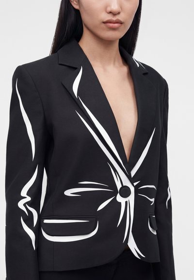 Γυναίκα με μαύρο σακάκι με λευκά αφηρημένα σχεδιαγράμματα, κλείσιμο με ένα κουμπί και τσέπες με καπάκι, εμφανιζόμενη από το στήθος και πάνω.