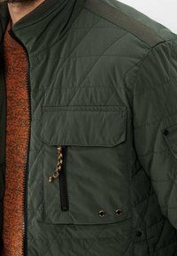 Grøn quiltet jakke med lynlås foran, brystlomme og ribbet krave. Har tekstureret stof og kontrasterende orange inderfor.