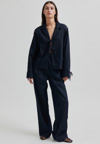 Îmbrăcăminte din denim închis, care include o cămașă lejeră cu un nod în față și mâneci lungi, asociată cu pantaloni cu picioare largi. Ambele piese au detalii minime.