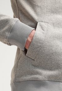 Felpa grigia chiaro con una texture accogliente, polsini a coste e una tasca frontale con una finitura liscia. Design semplice e minimalista.