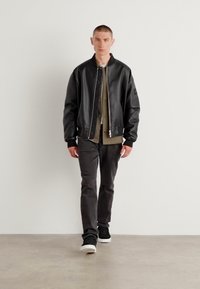 Zwart leren bomberjack met geribde manchetten, laag over een gedempt shirt, gecombineerd met donkere broek en zwarte sneakers. Model staat tegen een neutrale achtergrond.