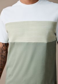 Rövid ujjú póló fehér felső résszel, strukturált világoszöld középső csíkkal és egyszínű zöld alsó résszel; pamut anyagból.