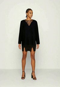 Vestido preto de manga longa com detalhe de decote em V em renda e saia justa com fendas laterais, comsapatos de salto alto pretos e atados.