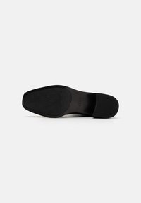 Semelle de chaussure noire avec un motif texturé, présentant une finition lisse et une discrète empreinte de logo, conçue pour la durabilité et l'adhérence.