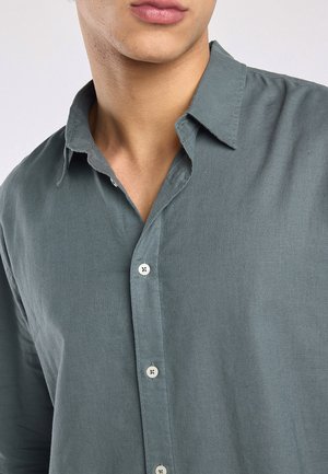 Close-up van een persoon die een grijs overhemd met knopen draagt en een open kraag, waarbij het onderste deel van het gezicht en de nek zichtbaar is.