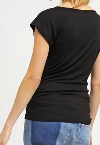 Žena v přiléhavém černém tričku s krátkým rukávem a dvoubarevných modrých džínech, zobrazená zezadu, s mírně vztyčenou rukou.