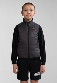 Gilet en duvet gris avec un col haut, fermeture éclair, poches latérales et un patch logo, superposé sur une chemise à manches longues noire et un short.