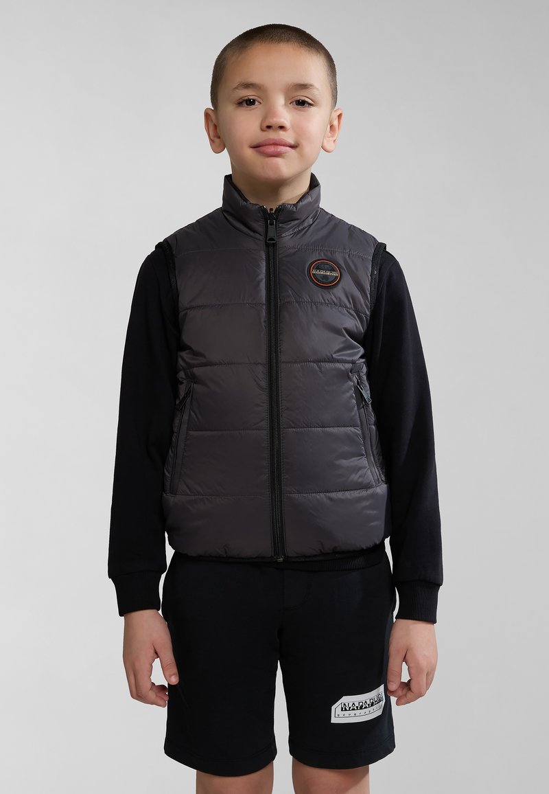 Gilet en duvet gris avec un col haut, fermeture éclair, poches latérales et un patch logo, superposé sur une chemise à manches longues noire et un short.