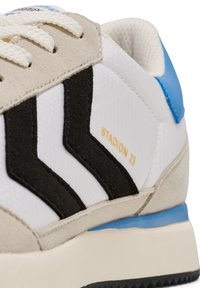 Vit sneaker med svarta mocka-ränder, ljusgrå mocka nära snörningen, en blå detalj på hälen och en texturerad yta.