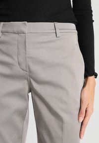 Žena v světle šedých tailored kalhotách a černém žebrovaném topu s dlouhým rukávem, ruka opřená o stehno.