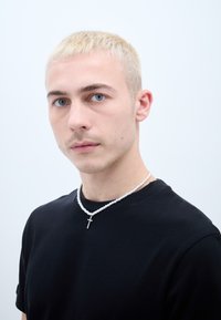 Balta pērļu kaklarota ar mazu sudraba krustu, uzvilkta virs melna T-kreklu. Modelim ir īsi, gaiši blondi mati un zili acu.