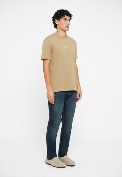 Tricou din bumbac bej, cu decolteu rotund și logo brodat, asociat cu blugi negri și mocasini crem pe un model.