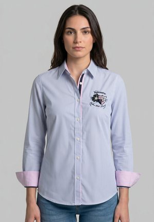 Mujer que lleva una camisa de rayas finas azul claro con puños rosas y un escudo bordado en el pecho izquierdo, de pie frente a un fondo gris.