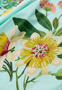 Bloemenstof met grote gele en witte bloemen met rode accenten op een lichtblauwe achtergrond, aangevuld met groene bladeren.