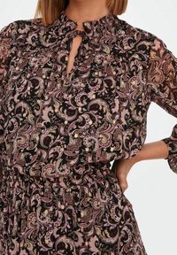Blouse noire à motif floral avec tissu transparent, col à volants, détail œillet et taille élastique. Manches longues avec poignets élastiques.