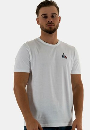 Camiseta de algodón blanca con mangas cortas, cuello redondo y un pequeño logo bordado en el lado izquierdo del pecho. Textura suave, corte clásico.
