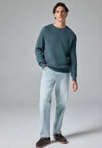 Pull à côtes teal foncé, jeans bleu clair et chaussures à lacets marron avec une semelle épaisse. Le mannequin se tient les mains dans les poches devant un fond neutre.