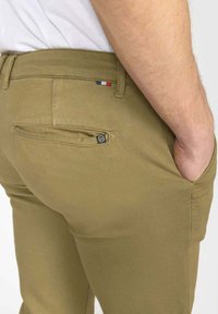 Beige chino's gedragen door een persoon met hand in de voorzak, waarbij de achterzak zichtbaar is met een klein zwart etiket en een klein detail van rode, witte en blauwe stof.