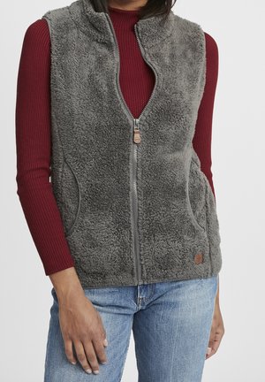 Femme portant un gilet en polaire gris à fermeture éclair par-dessus un t-shirt rouge côtelé à manches longues et un jean bleu, debout avec une main près de la hanche.