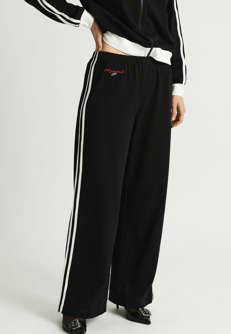 Persoană purtând pantaloni sport negri cu picioare largi și dungi laterale albe, cu text brodat roșu, asortați cu pantofi eleganți negri și o jachetă asortată.