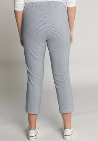 Persoon gekleed in cropped slim-fit broek met zwart-wit geblokt patroon en witte sneakers, staand tegen een effen achtergrond.