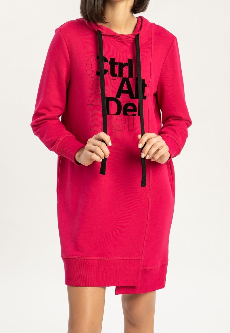 Kobieta w jasnoróżowej sukience z kapturem, z czarnymi sznurkami i nakładającym się napisem "Ctrl Alt Del" z przodu.