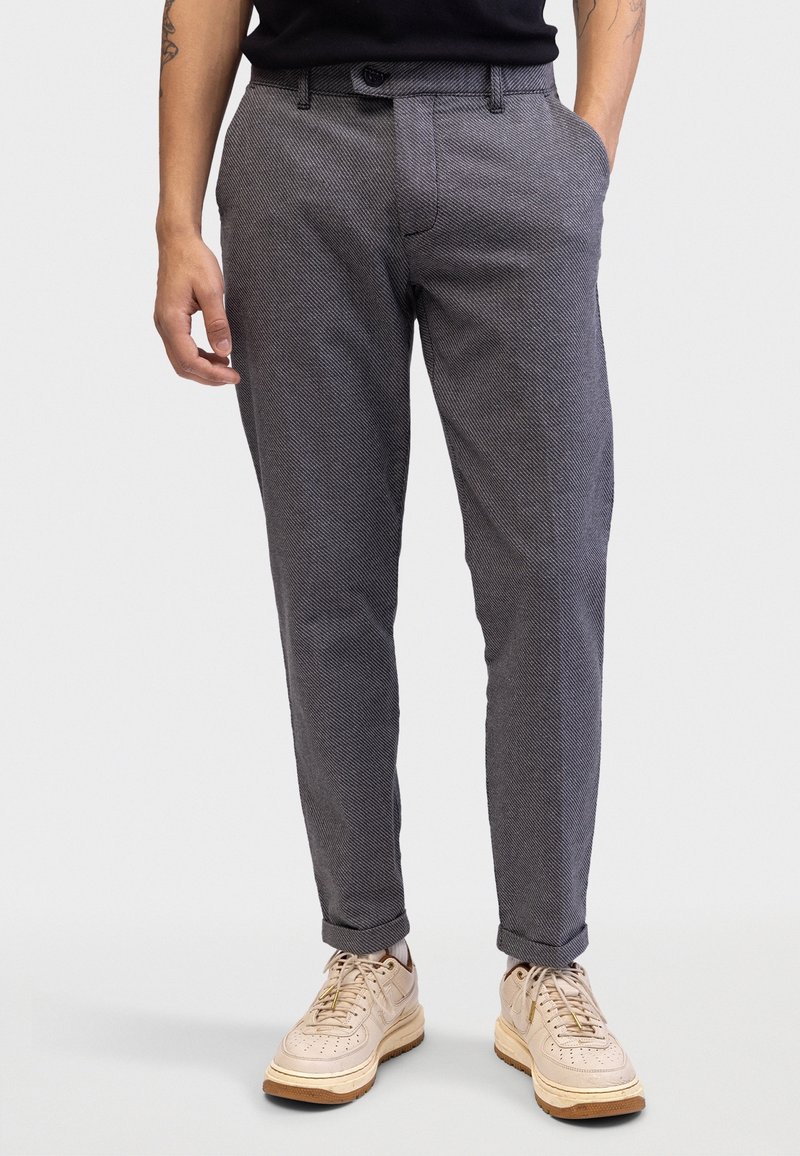 Homme portant un pantalon gris texturé et fuselé avec des ourlets retroussés, des baskets beiges, une chemise noire, debout avec une main dans la poche.