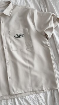 Lyhythihainen beige paita, joka on valmistettu sileästä kankaasta. Paidassa on rintatasku mustalla logolla ja napit edessä.