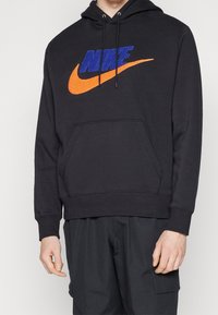 Sudadera negra con un bolsillo tipo canguro en el frente, que presenta un gran logo de Nike en azul y naranja. La tela parece suave y tiene un ajuste relajado.