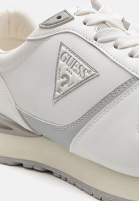 Zapatilla blanca con un acabado de piel lisa, acentos grises, entresuela texturizada y un logo metálico triangular en el lateral.