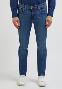 Jeans de ganga azul com um corte justo, cinco bolsos e costura em contraste. Combinados com ténis brancos, apresentando um estilo casual.