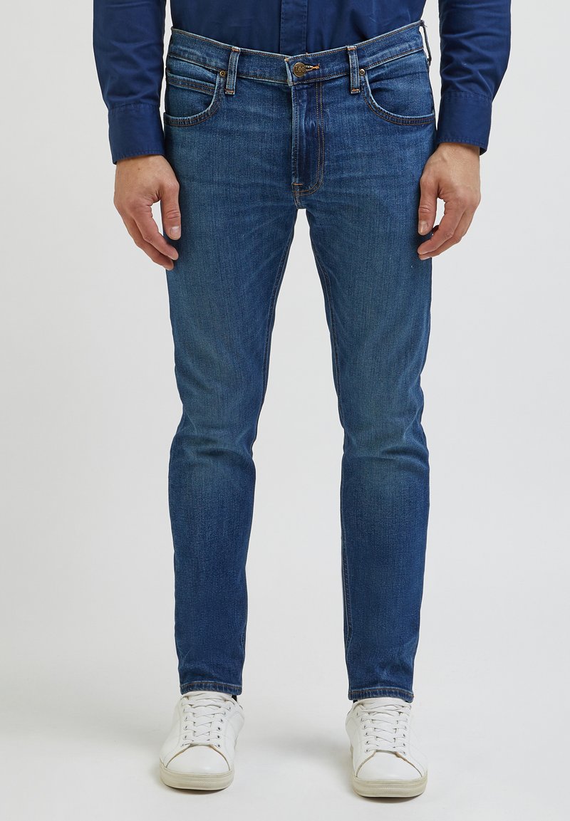 Jeans de ganga azul com um corte justo, cinco bolsos e costura em contraste. Combinados com ténis brancos, apresentando um estilo casual.