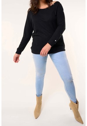 Top negro de manga larga con diseño off-shoulder y detalles de botones. Combinado con vaqueros ajustados azul claro y botas de tobillo puntiagudas beige.