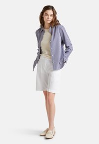 Camicia a righe blu e bianca con bottoni indossata sopra un maglione beige, abbinata a pantaloni corti bianchi fino al ginocchio e scarpe slip-on di colore chiaro.