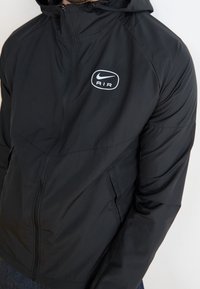 Černá bunda Nike se zipem vpředu, kapucí a logem. Má hladkou texturu, přiléhavý střih a boční kapsy pro praktičnost.