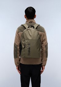 Olivgrön ryggsäck med en strukturerad yta, justerbara remmar och en svart logotyp på framsidan. Buren över en beige fleecejacka.
