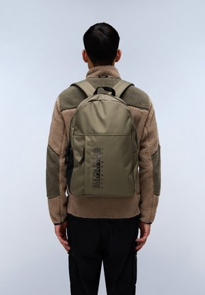 Mochila de color verde oliva con una superficie texturizada, correas ajustables y un logo negro en la parte frontal. Llevada sobre una chaqueta de forro polar beige.