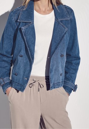 Femme portant une veste en jean bleue par-dessus un t-shirt blanc et un pantalon beige à cordon, les mains dans les poches, sur un fond uni.