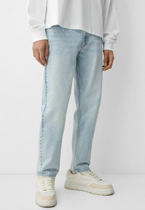 Personne portant un jean droit bleu clair, des baskets blanches et un haut blanc à manches longues ample, debout devant un fond uni.