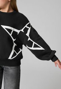Svart sweatshirt med vit grafisk stjärndesign. Har nedhasade axlar, ribbade muddar och nederkant, i en mjuk tygtextur. Bärs med grå jeans.