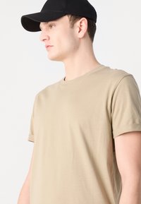 Tricou bej cu guler rotund, cu mâneci scurte, răsucite, realizat dintr-un material neted. Purta o șapcă de baseball neagră, arătând profilul lateral.
