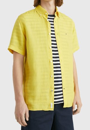 Mann trägt ein aufgeknöpftes gelbes Kurzarmhemd über einem schwarz-weiß gestreiften T-Shirt, kombiniert mit dunklen Shorts, vor einem einfarbigen Hintergrund.